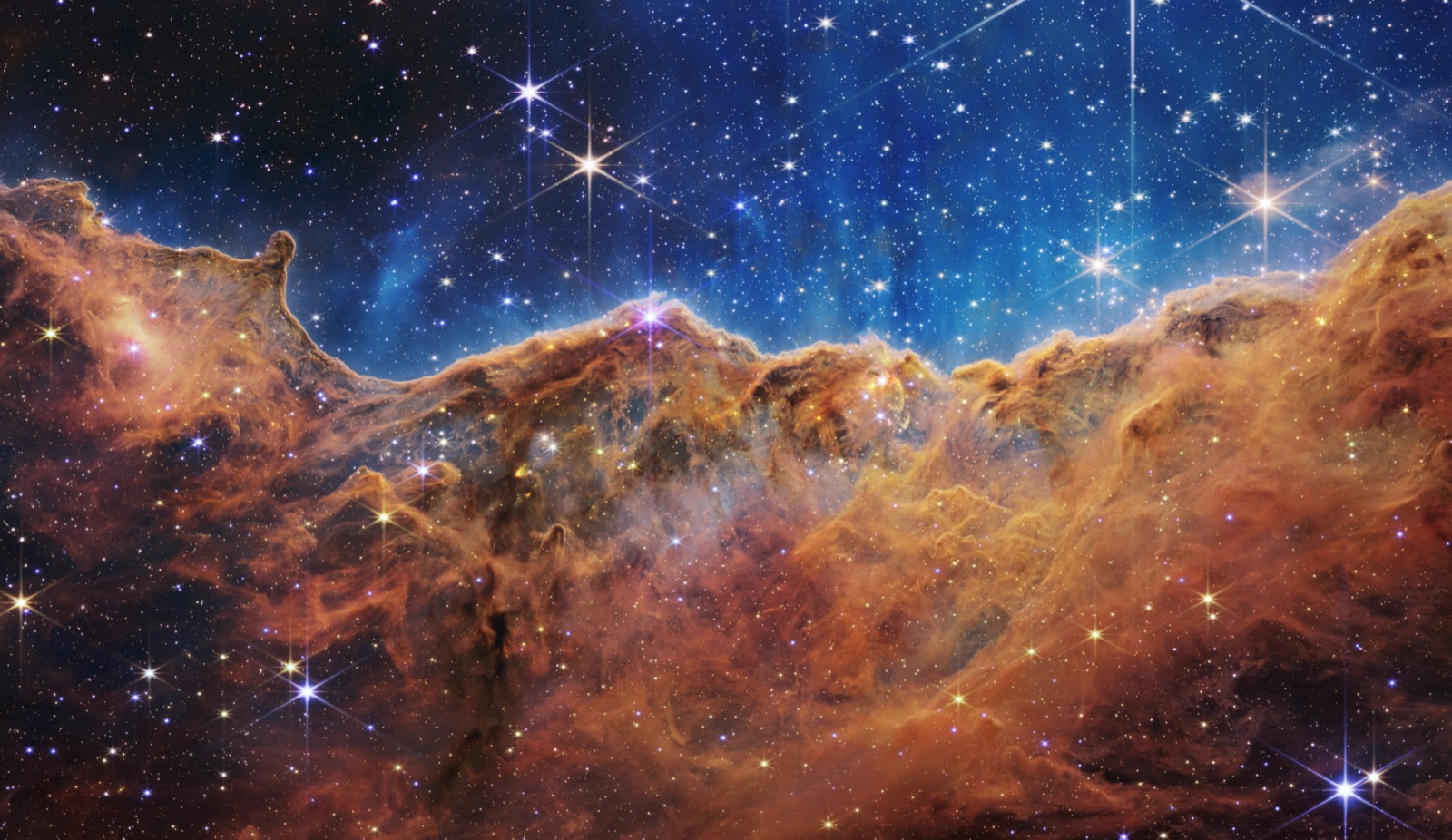 Webb image of the Cosmic Cliffs in the Carina Nebula, where the life cycle of a star begins: an orange ridge of gas and dust beneath a blue starry sky