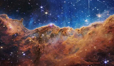 The Cosmic Cliffs in the Carina Nebula imaged by the James Webb Space Telescope: a glowing ridge of gas and dust under a star-filled blue sky.